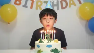 Boy Blowing Out Candles On A Dinosaur Cake At His Birthday Party